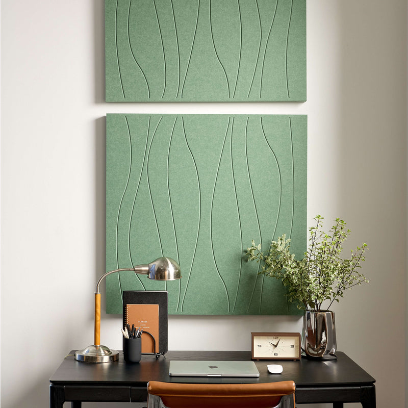 A large rectangular wall art panel with a sage green textured background and thin black wavy line pattern, shown hanging on a light wall in a styled interior with a desk lamp, notebook, plant, and small clock.