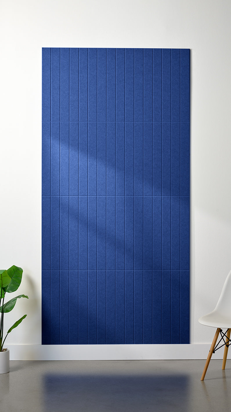 Blue Felt Right 4’ x 8’ PET felt panel with straight vertical lines engraved into the material against a white wall. A plant and chair are in the foreground.