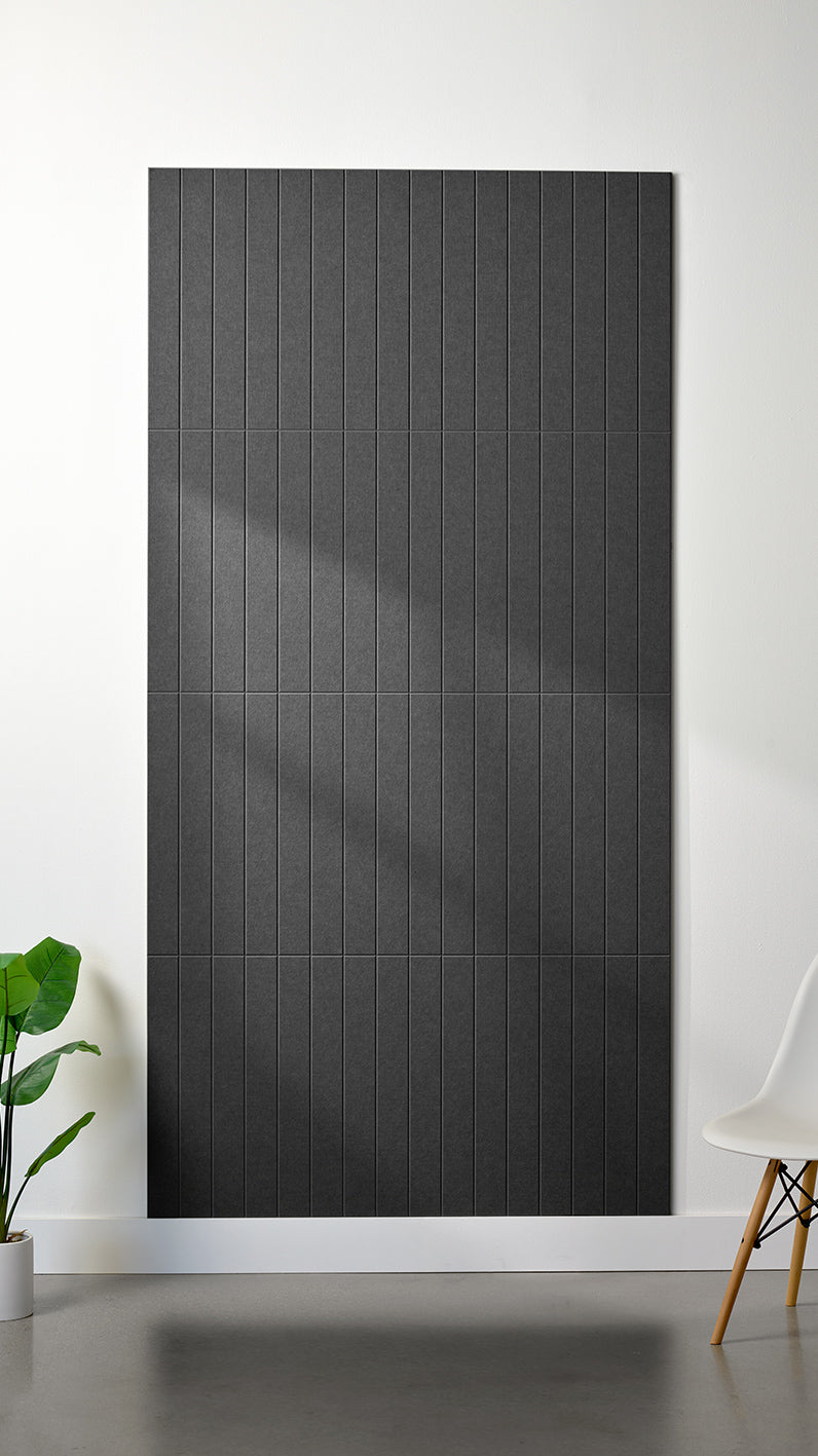 Dark grey Felt Right 4’ x 8’ PET felt panel with straight vertical lines engraved into the material against a white wall. A plant and chair are in the foreground.