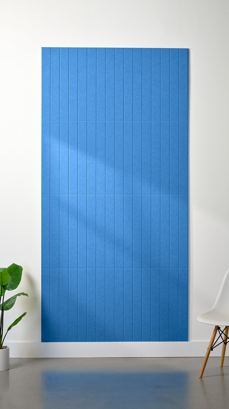 Bright blue Felt Right 4’ x 8’ PET felt panel with straight vertical lines engraved into the material against a white wall. A plant and chair are in the foreground.