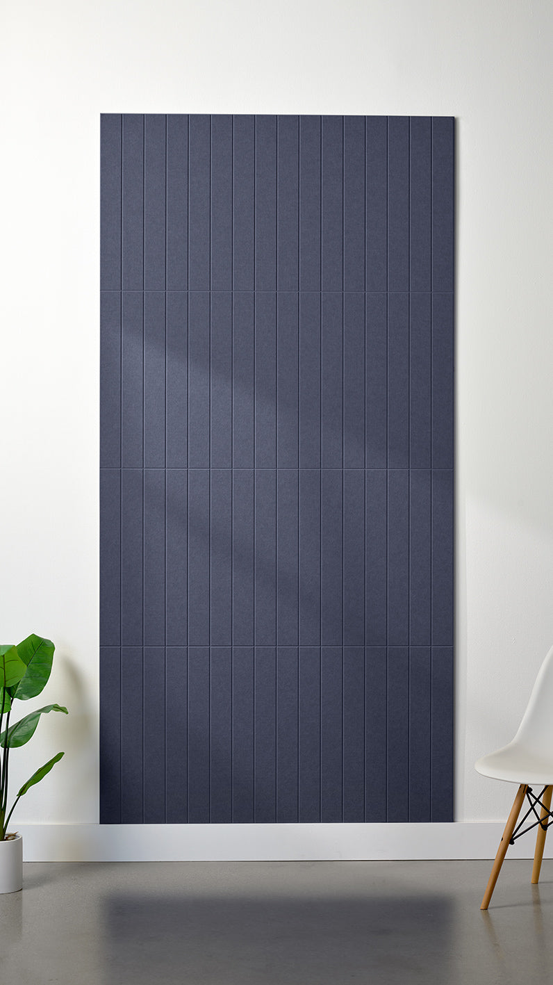 Indigo Felt Right 4’ x 8’ PET felt panel with straight vertical lines engraved into the material against a white wall. A plant and chair are in the foreground.