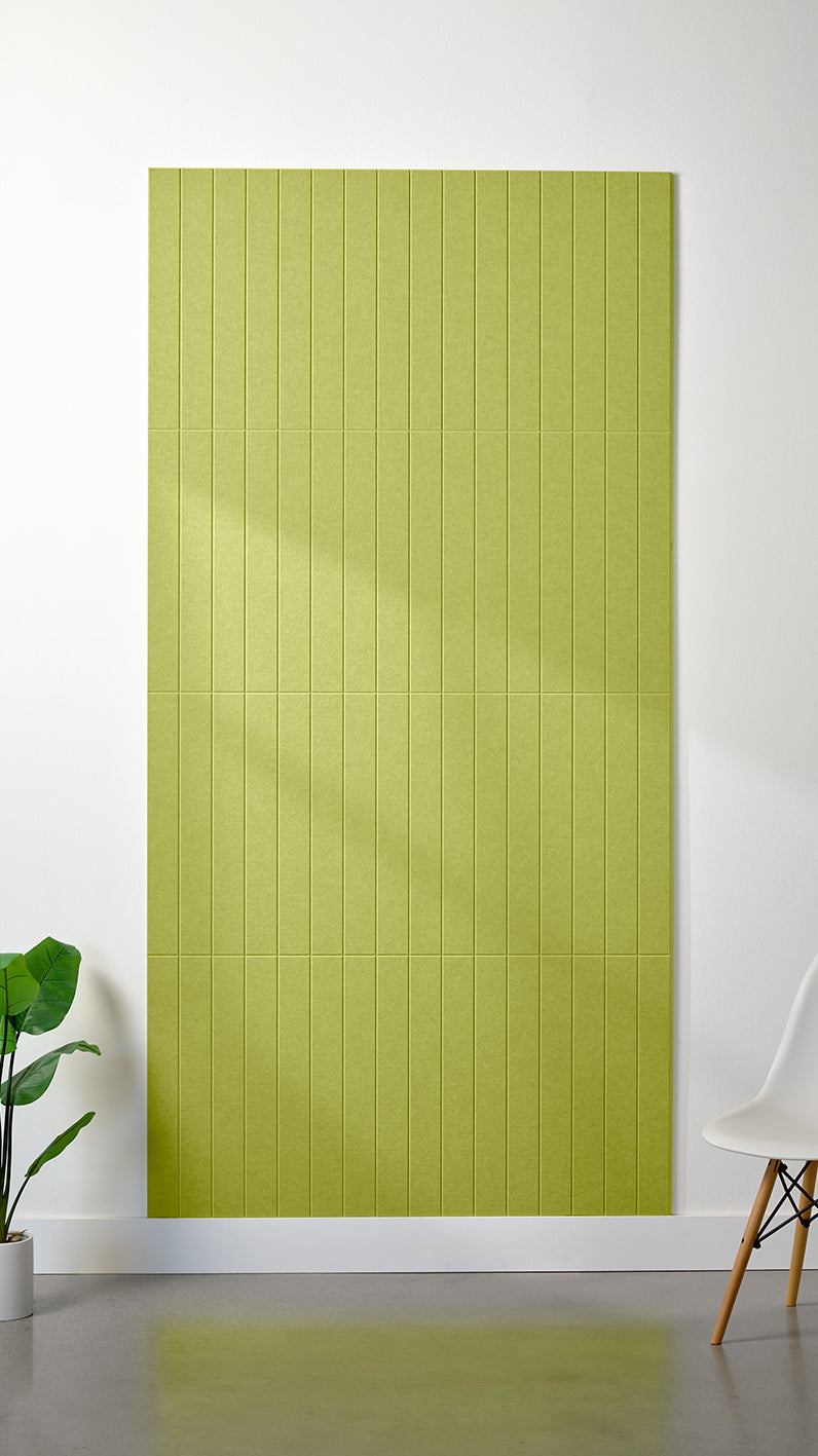 Bright green Felt Right 4’ x 8’ PET felt panel with straight vertical lines engraved into the material against a white wall. A plant and chair are in the foreground.