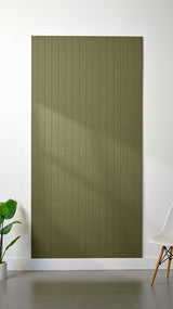 Olive green Felt Right 4’ x 8’ PET felt panel with straight vertical lines engraved into the material against a white wall. A plant and chair are in the foreground.