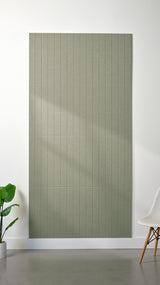 Sage green Felt Right 4’ x 8’ PET felt panel with straight vertical lines engraved into the material against a white wall. A plant and chair are in the foreground.