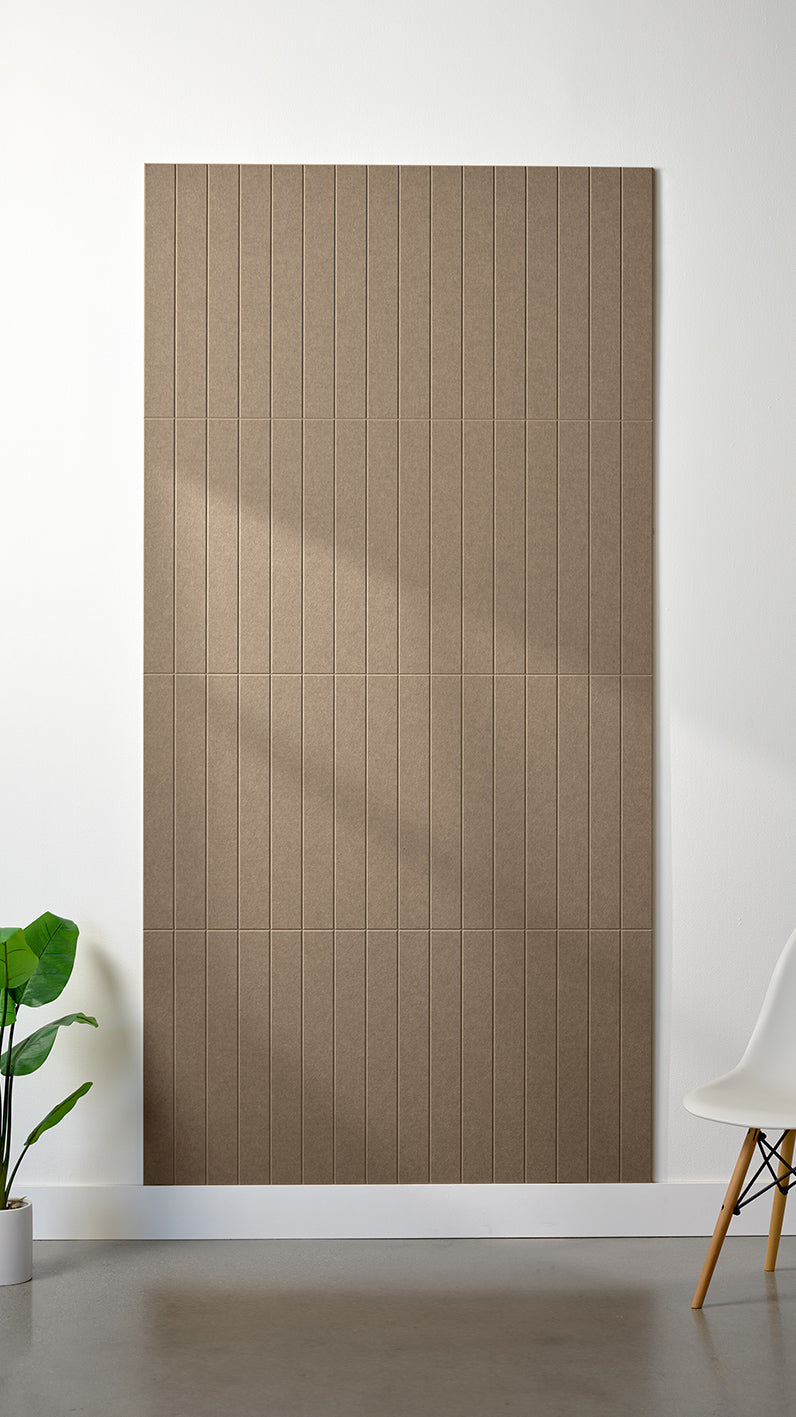 Light brown Felt Right 4’ x 8’ PET felt panel with straight vertical lines engraved into the material against a white wall. A plant and chair are in the foreground.