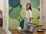 Woman standing in a room with green and blue circular wall panels, using a laptop on a table.