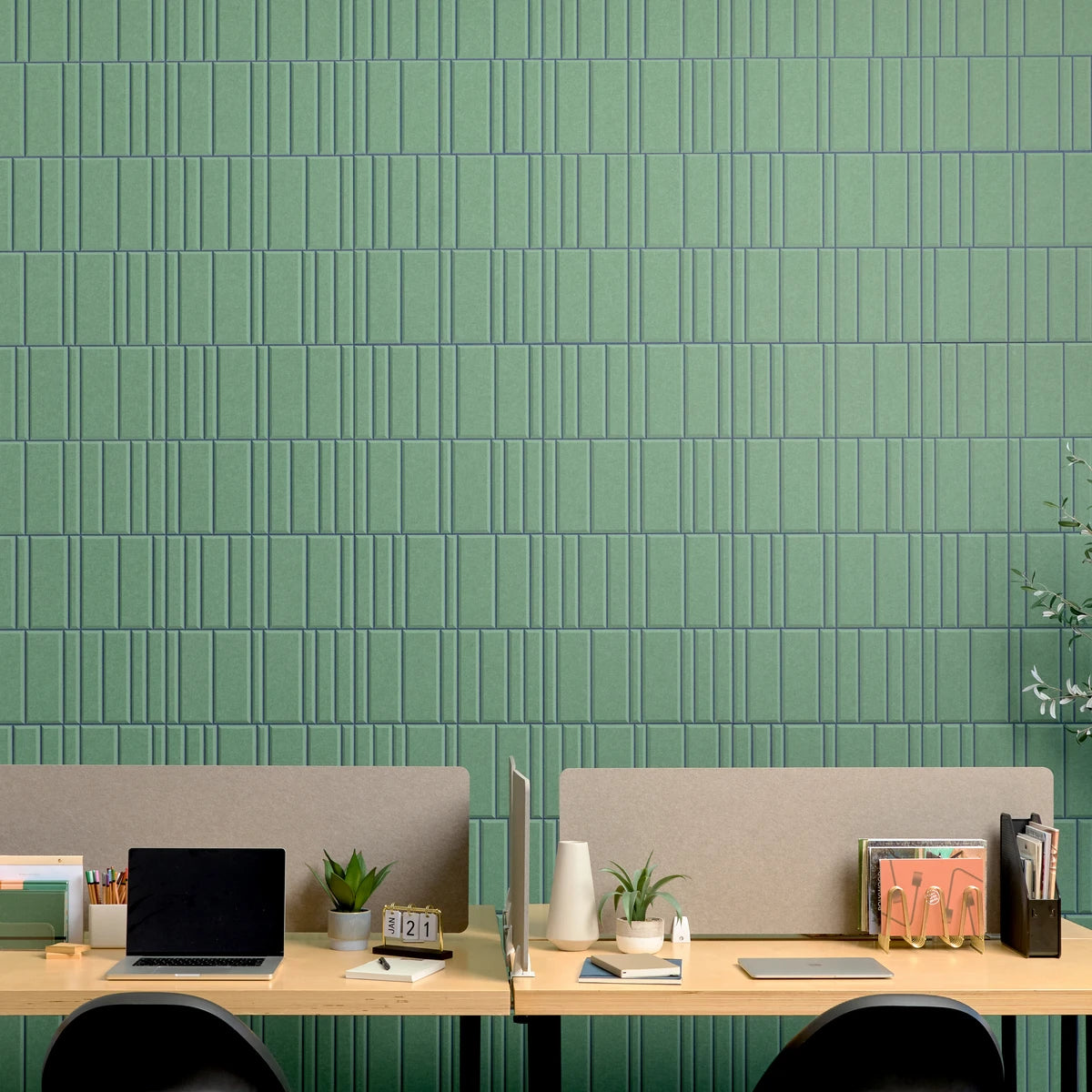 A modern office setup with two light wood desks, beige felt dividers, and black chairs in front of a prominent green textured acoustic wall with vertical rectangular tiles.