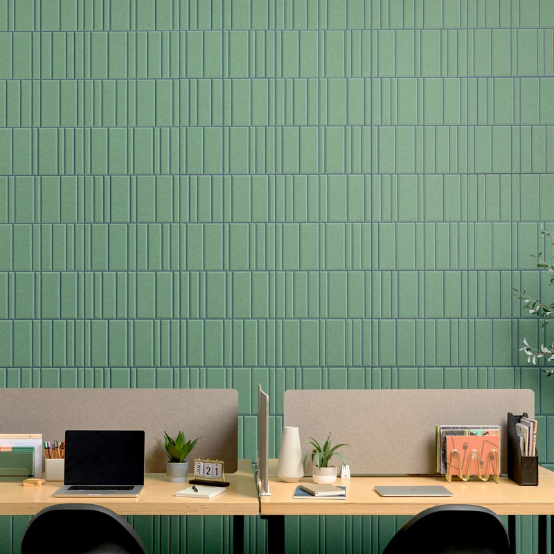 A modern office setup with two light wood desks, beige felt dividers, and black chairs in front of a prominent green textured acoustic wall with vertical rectangular tiles.
