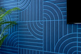 Blue tiles with layered geometric arc and line engraving pattern installed on wall with green plant visible.