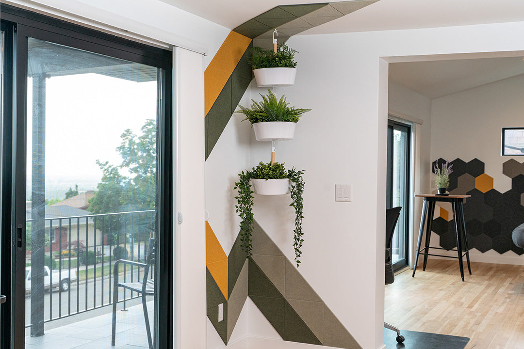 A Felt Right wall art design in shades of yellow and green felt tiles in a lightning like stripe on a white wall in a room next to a sliding glass door and three hanging house plants.