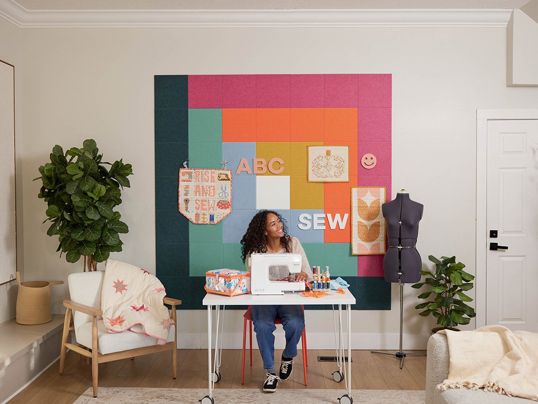 Felt right wall art in a craft room or sewing room in shades of green, blue, pink, and orange felt square tiles in a quilt pattern called log cabin with a woman working at a sewing machine and a chair with a quilt in the corner.
