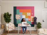 Felt right wall art in a craft room or sewing room in shades of green, blue, pink, and orange felt square tiles in a quilt pattern called log cabin with a woman working at a sewing machine and a chair with a quilt in the corner.