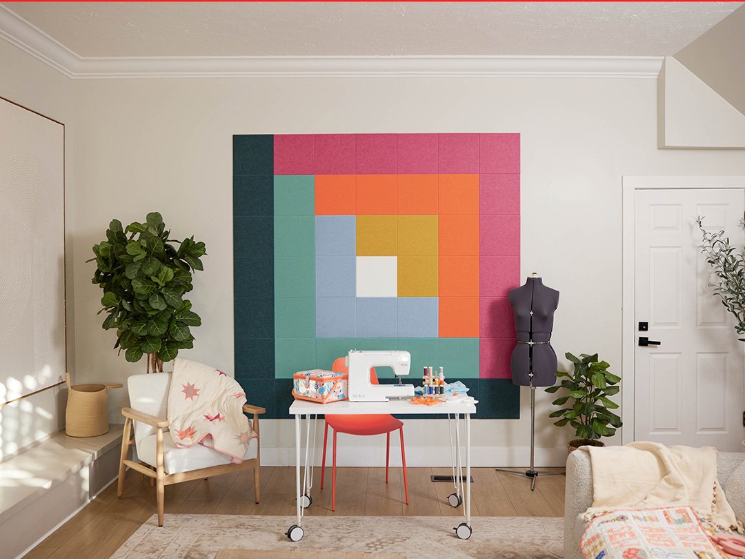 Felt right wall art in a craft room or sewing room in shades of green, blue, pink, and orange felt square tiles in a quilt pattern called log cabin behind a desk with a sewing machines and chair in the corner with a quilt on it.