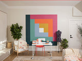 Felt right wall art in a craft room or sewing room in shades of green, blue, pink, and orange felt square tiles in a quilt pattern called log cabin behind a desk with a sewing machines and chair in the corner with a quilt on it.