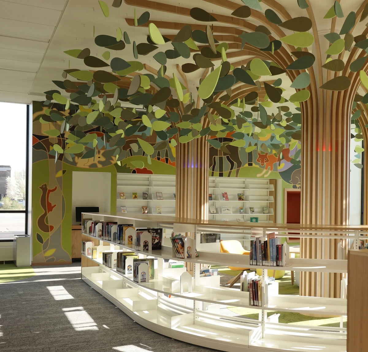 A modern library or study interior with curved white bookshelves, tall slatted wood columns, and a large hanging leaf canopy suspended from the ceiling. Colorful abstract wall murals are visible in the background.