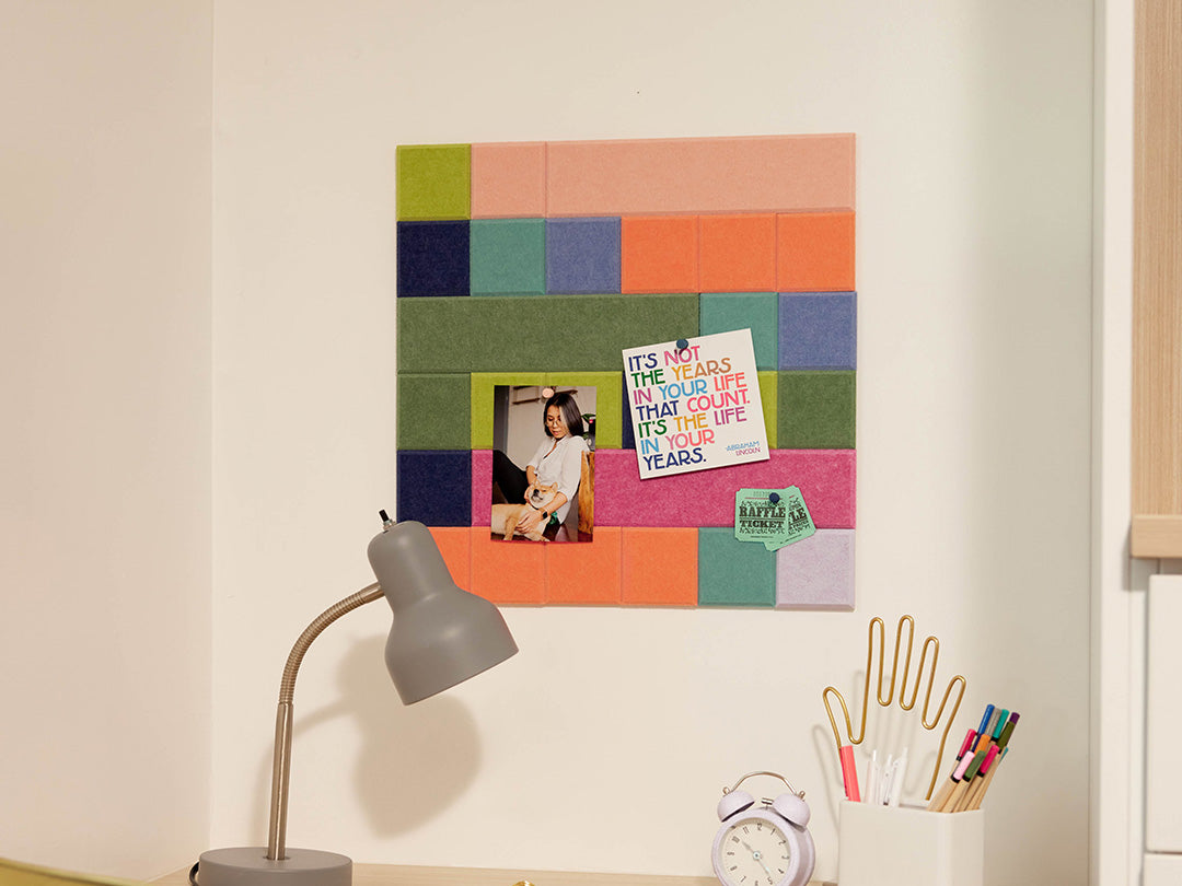 Colorful square bulletin board on a wall with a lamp, clock, and stationery items below.
