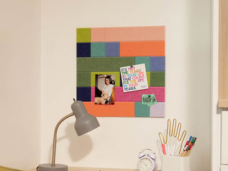 Colorful square bulletin board on a wall with a lamp, clock, and stationery items below.