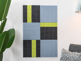 Felt Right wall art design on a white wall using blue, grey, and green felt tiles next to a house plant and a chair.
