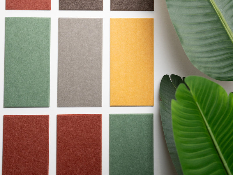 A Felt Right wall design in array of solid color sample tiles arranged in a grid pattern on a wall, with a variety of colors including greens, yellows, reds, and grays, and a green plant in the bottom right corner.