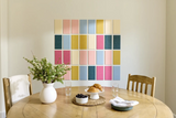 A styled dining room scene with a round wooden table, chairs, a white vase with greenery, and a colorful grid wall hanging made of pastel and muted colored rectangular panels behind the table.