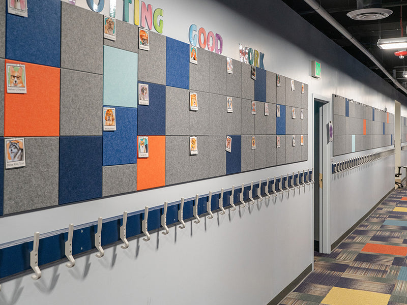 A Felt Right wall art design in shades of grey, blue, and orange square felt tiles with a pinnable welcome banner on a wall above a coat rack in a classroom.