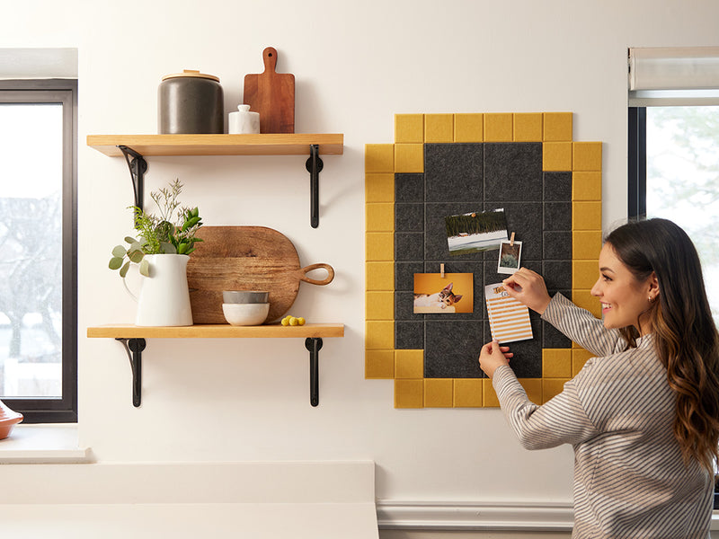 Felt Right wall art tiles in shades of black and yellow creating a framed look with a woman pinning cards, photos, and to-do lists,  on a wall in a kitchen by shelves and windows