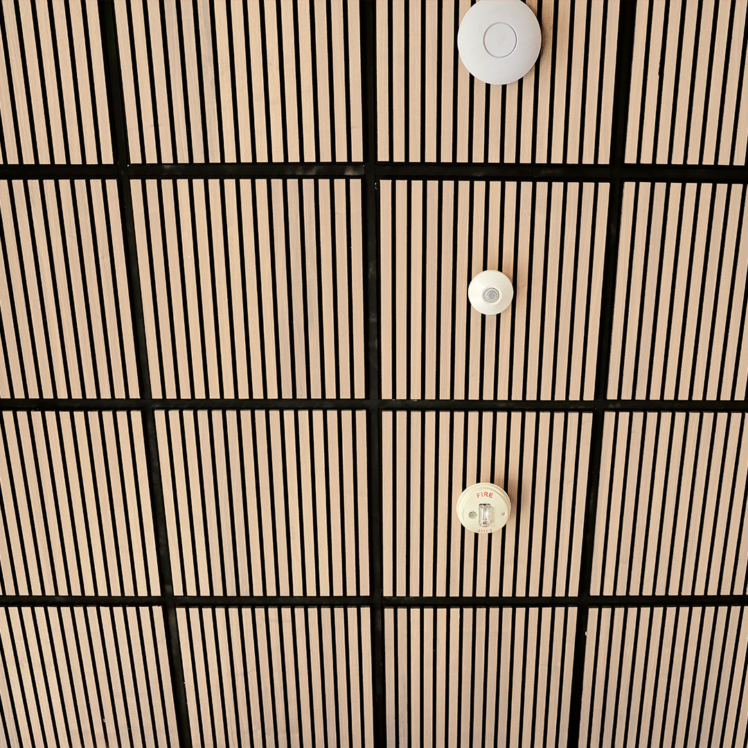 A close-up of a modern ceiling with a grid pattern of light wood slats and black grout lines, featuring three white circular ceiling-mounted sensors or diffusers.