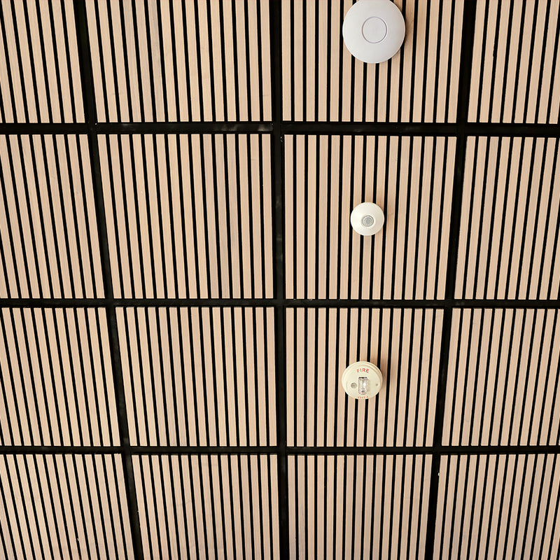 A close-up of a modern ceiling with a grid pattern of light wood slats and black grout lines, featuring three white circular ceiling-mounted sensors or diffusers.