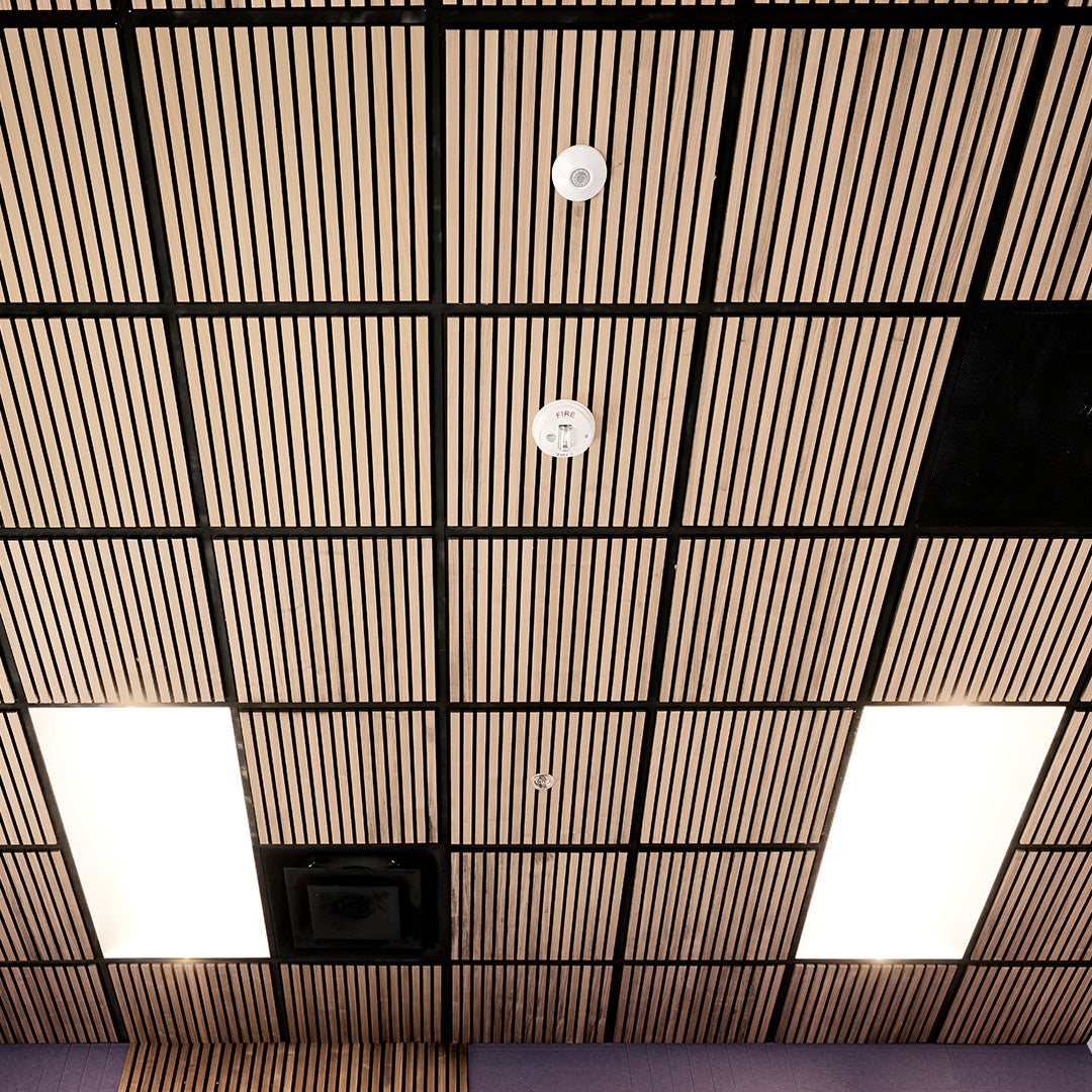 Wood slat ceiling panels arranged in a grid with black metal grid lines, recessed square lights, smoke detectors, and a ventilation opening.