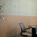 A modern interior corner with a light wood slatted wall paneling, a pale green patterned upper wall, a black office chair with a beige seat, and a dark round table holding books and a glass.