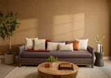 A modern living room scene featuring a gray fabric sofa with multiple decorative throw pillows, set against a warm wood slat wall. A round wooden coffee table sits in front of the sofa, and potted plants and side tables are arranged around the room.