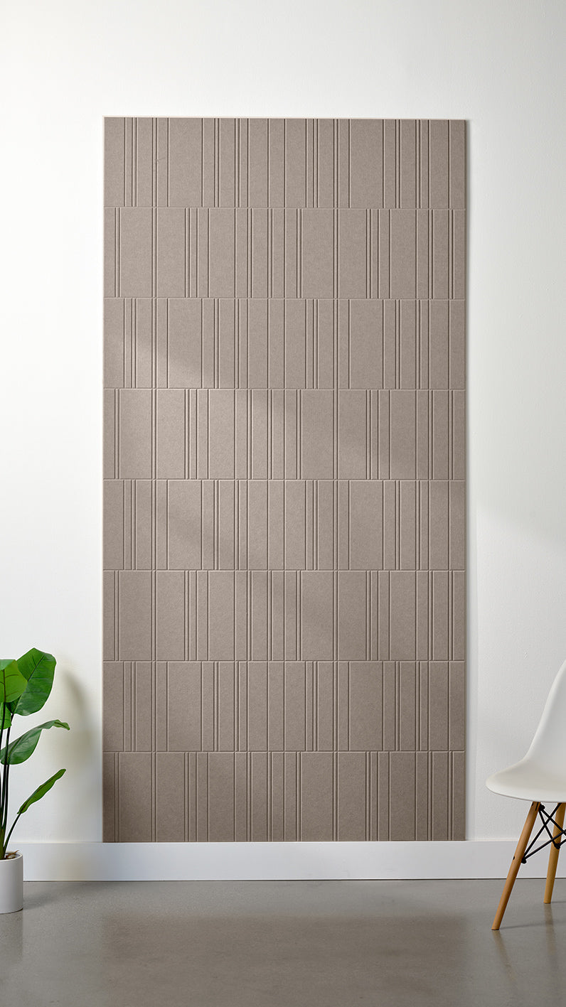 Greyish beige Felt Right 4’ x 8’ PET felt panel with staggered lines engraved into the material against a white wall. A plant and chair are in the foreground.