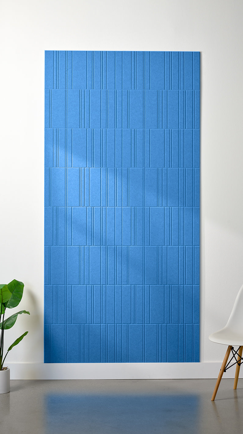 Bright blue Felt Right 4’ x 8’ PET felt panel with staggered lines engraved into the material against a white wall. A plant and chair are in the foreground.