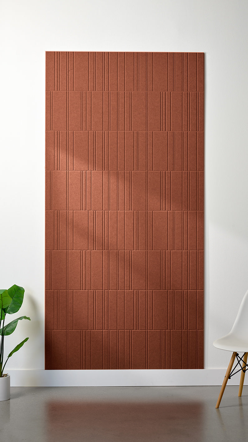 Burnt orange Felt Right 4’ x 8’ PET felt panel with staggered lines engraved into the material against a white wall. A plant and chair are in the foreground.