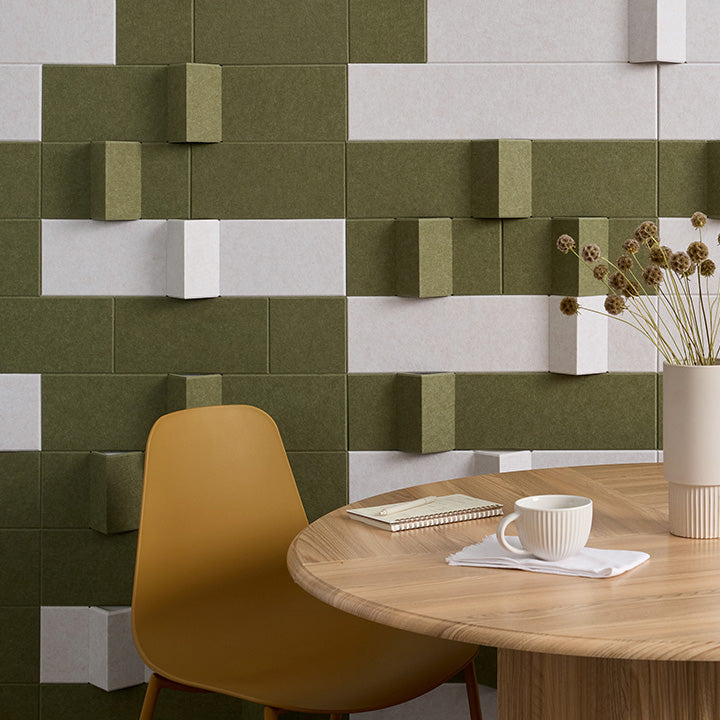 Olive green and off-white 3D geometric wall panels arranged in a modern checkered pattern behind a round wooden dining table, a mustard yellow chair, and a white vase with dried flowers.