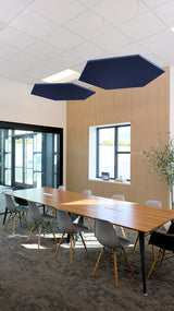 Two hexagon-shaped navy baffles with wood slat backing suspended above a conference table in an office setting.