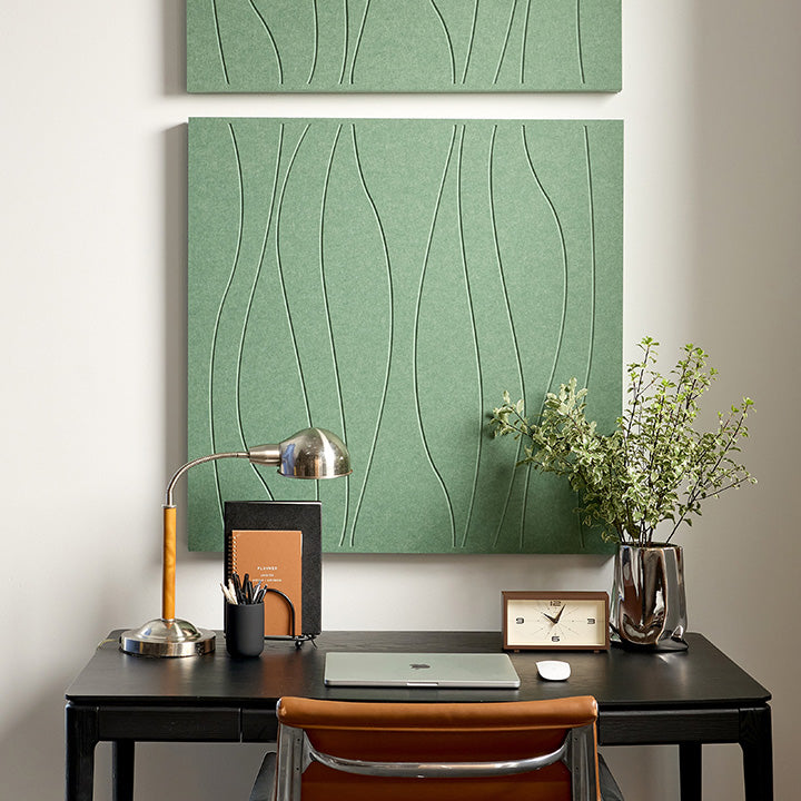 Two green textured acoustic wall art panels with raised wavy lines hang above a black desk in a styled home office setting. The desk includes a silver desk lamp, notebook, cup, laptop, mouse, small clock, and a vase with greenery.