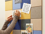 A Felt Right wall art design using squares to form a pattern in shades of blue, cream, grey, and yellow felt tiles and a woman oinning art or cards to the tiles