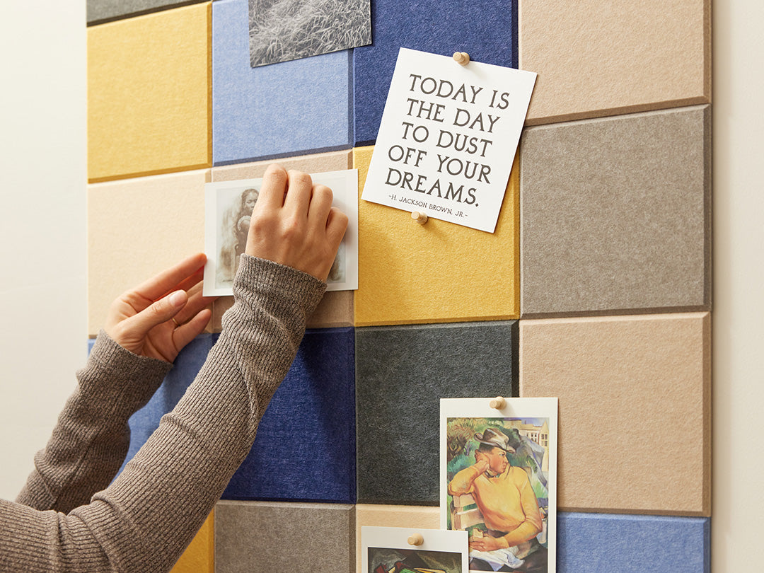 A Felt Right wall art design using squares to form a pattern in shades of blue, cream, grey, and yellow felt tiles and a woman oinning art or cards to the tiles