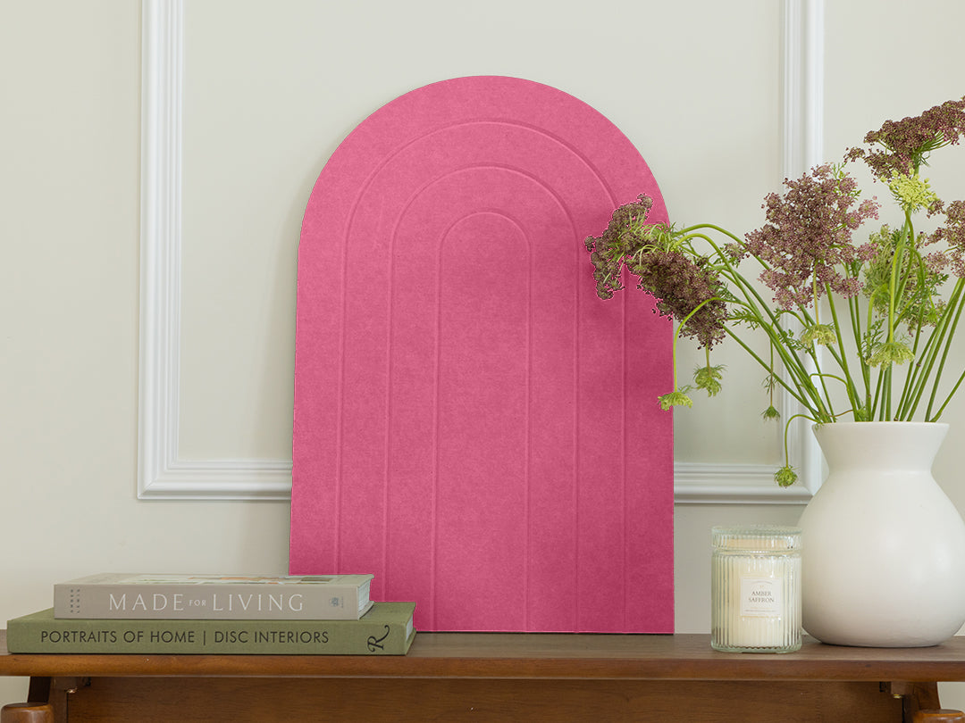 Pink arch-shaped wall art on a neutral wall with books and a vase of flowers on a wooden surface.