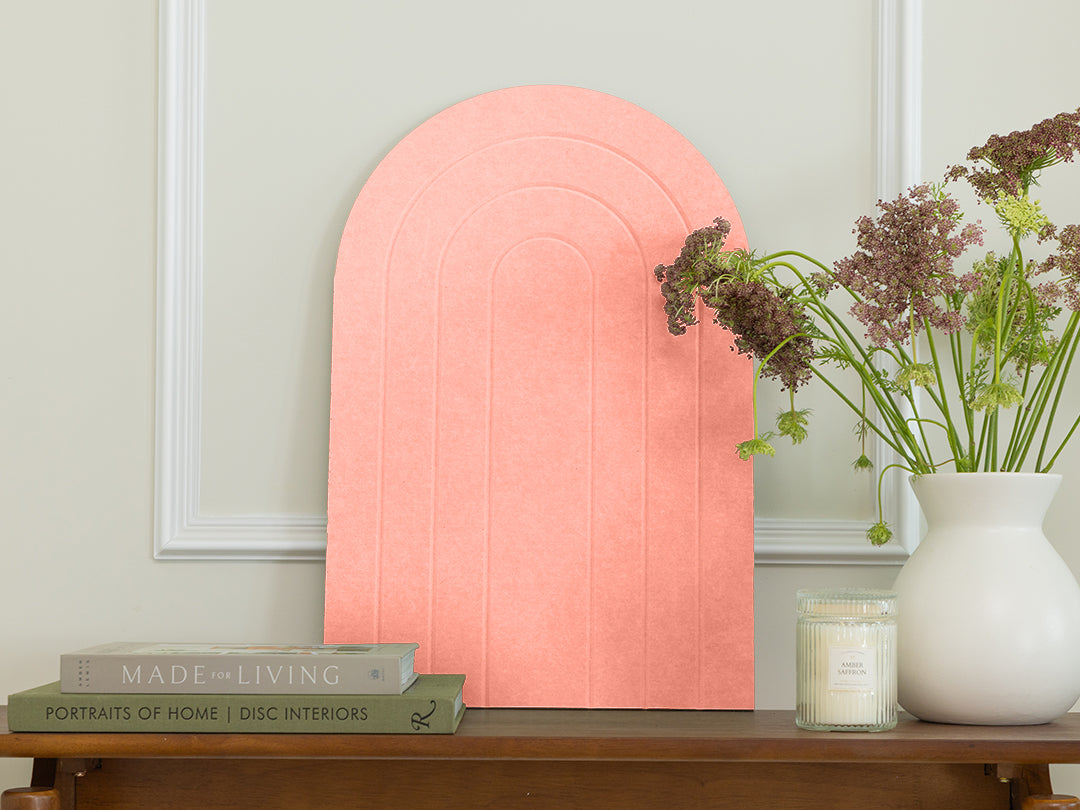 Pink arch-shaped wall art on a light gray wall with books and a vase on a wooden surface.