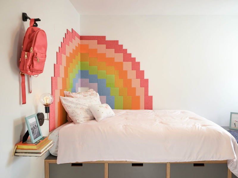 Felt Right wall art design using pixel square tiles in shades of the rainbow to create a pixel look of the rainbow on a corner of two walls in a bedroom next to a bed with a backpack hanging up.