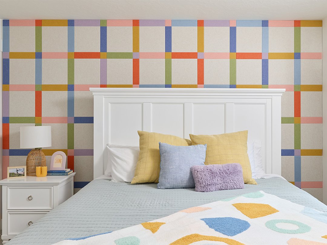 Felt Right wall art design in a bedroom with a plaid design using pink, orange, green, and blue felt tiles behind a bed and a night stand.