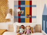 A Felt Right wall art design in shades of blue, yellow, and red felt tiles with photos pinned to it on a white wall in a children's room or playroom above a bed with two dolls on it.