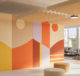 A modern interior space with a large abstract geometric wall mural in warm tones of orange, beige, yellow, and white. The room features layered wooden ceiling panels, pendant lights, and round upholstered ottomans in the foreground, with office desks visible through a large window on the right.