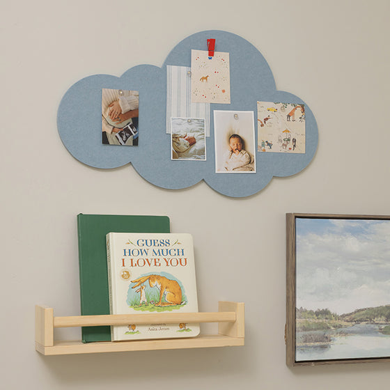 A light blue cloud-shaped wall organizer with photo clips and sample photos, mounted on a beige wall above a small wooden bookshelf holding children's books, with a framed landscape print to the right.