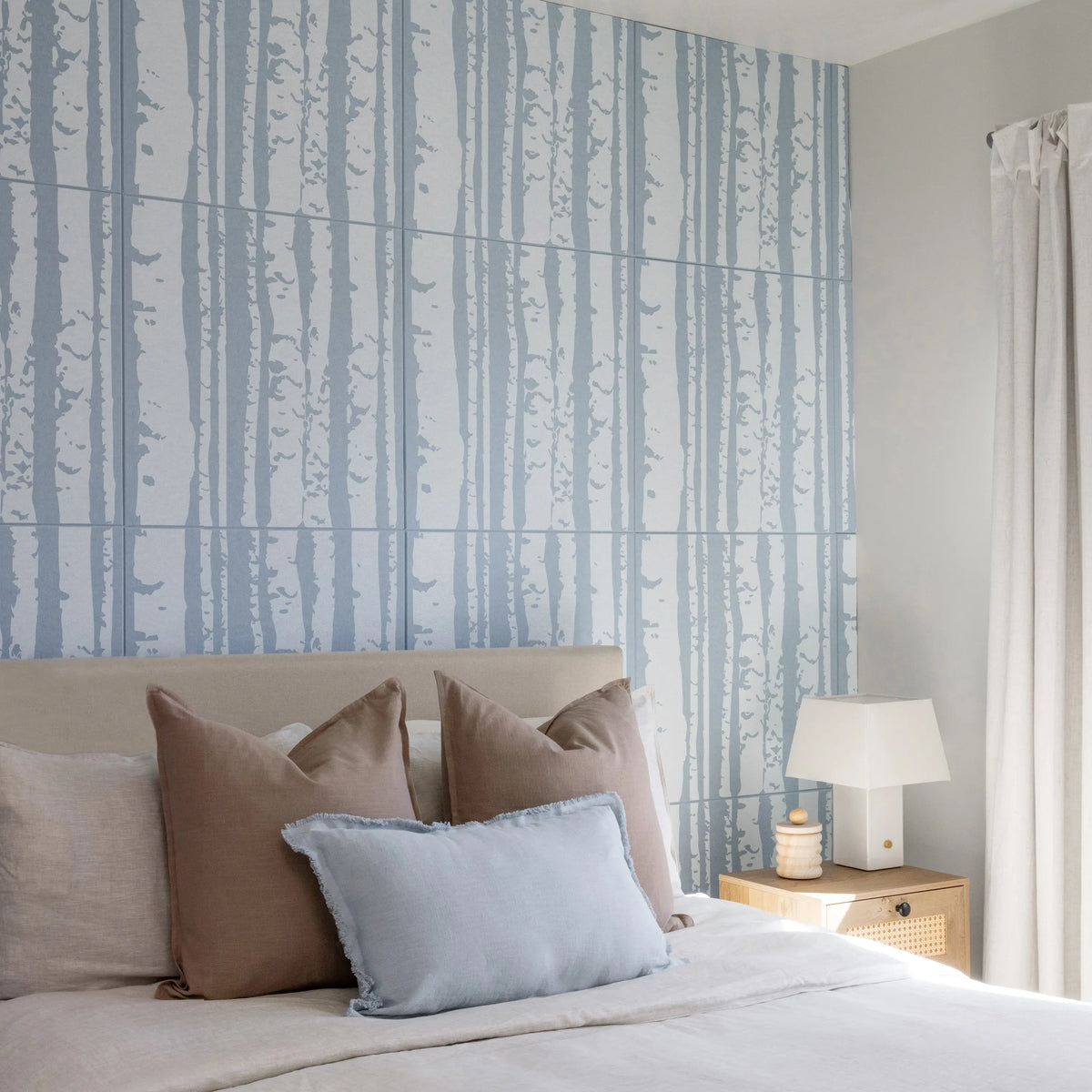 A styled bedroom featuring a large wall panel with a light blue and white vertical tree-bark pattern behind a bed with beige and light blue pillows. A potted green plant stands on the left, and a wooden nightstand with a white lamp is on the right.
