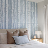 A styled bedroom featuring a large wall panel with a light blue and white vertical tree-bark pattern behind a bed with beige and light blue pillows. A potted green plant stands on the left, and a wooden nightstand with a white lamp is on the right.
