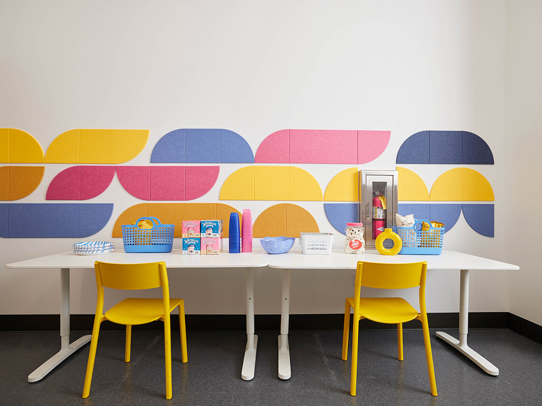 Break room with a white table and yellow chairs against a colorful geometric wall.