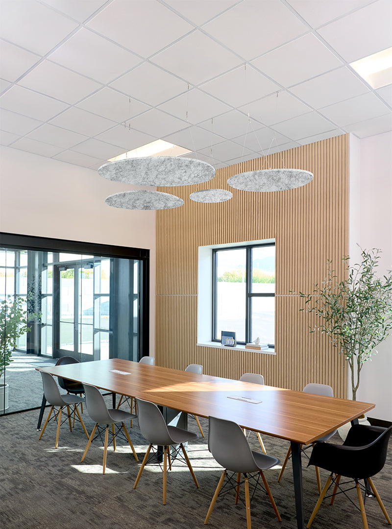 A modern conference room with a long wooden table, gray and black chairs, a slatted wood accent wall, large windows, and three oval textured pendant lights hanging from a white ceiling grid.